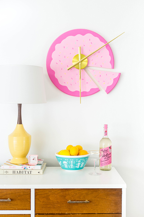 A pink wall clock shaped like a cake with a single detached piece hangs on a white wall over a buffet. On the buffet surface is a bowl of lemons, some decorative books, a table lamp, and a bottle of wine.